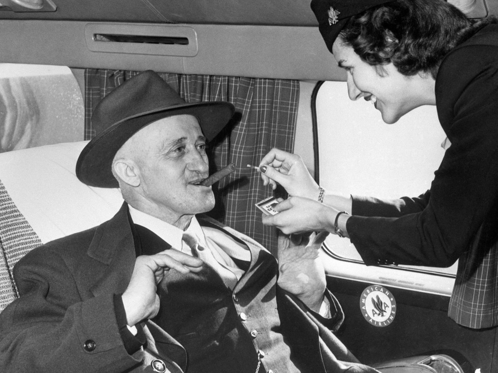 A stewardess helps a flyer light up a cigar midflight.